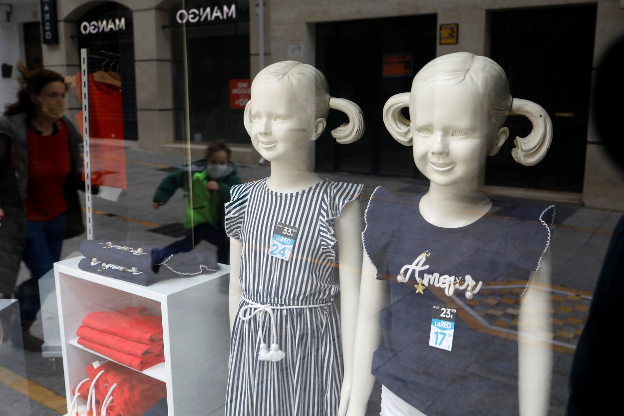 El reflejo de un niño y su madre, ambos con mascarilla, en el escaparate de una tienda de ropa infantil. REUTERS