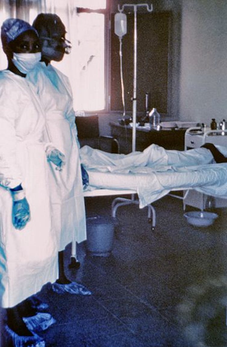 Fotografía de 1976, con dos enfermeras de pie junto a un paciente infectado por el virus del Ébola, que murió pocos días después debido a graves hemorragias internas. CDC, CC BY